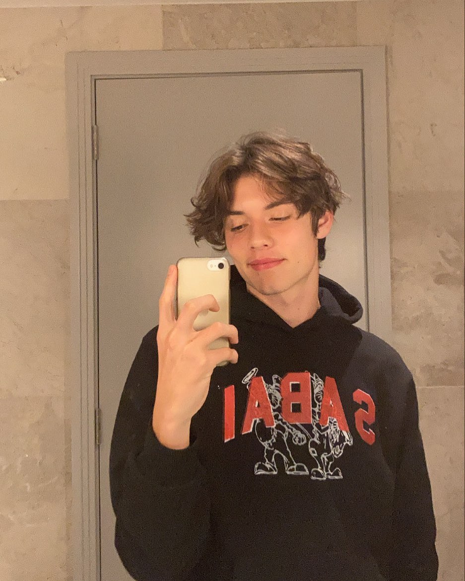 Louis Partridge Louis Partridge em selfie no espelho; ele usa um moletom preto com estampa e sorri levemente para foto que está sendo tirada com um celular branco