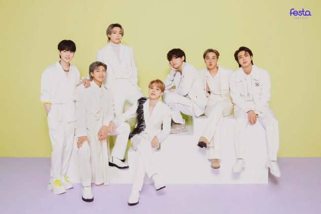 Grupo BTS posando para foto em fundo amarelo com chão em tom de lilás; todos os integrantes usam roupas brancas