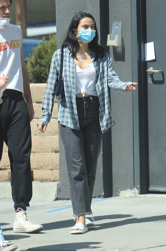 Camila Mendes caminhando pelas ruas de Los Angeles, usando máscara de proteção, camisa xadrez, calça jeans e chinelo branco.