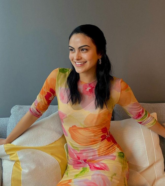 Camila mendes com vestido estampado Camila Mendes sentada em sofá branco, com os braços abertos, e expressão sorridente, ela usa vestido semitransparente com estampas nas cores rosa, laranja e verde.