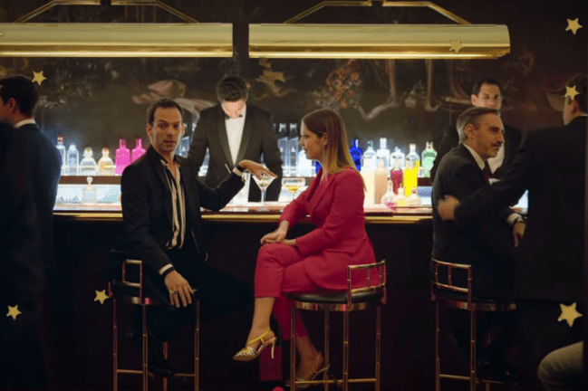 Conjunto rosa Caye Elite Personagem Cayetana do seriado Elite está sentada de lado, com um homem, ela usa conjunto monocromatico rosa pink e está em um bar.