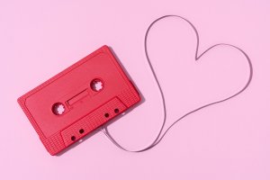 Uma fita cassete vermelha sobre um fundo rosa. A fitinha saindo dela forma um coração.