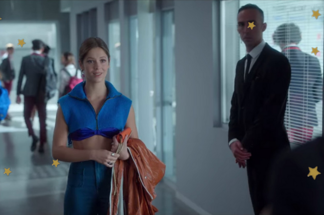 Look Caye quarta temporada Elite Personagem Cayetana do seriado Elite no corredor da escola carregando sua blusa marrom em uma das mãos.