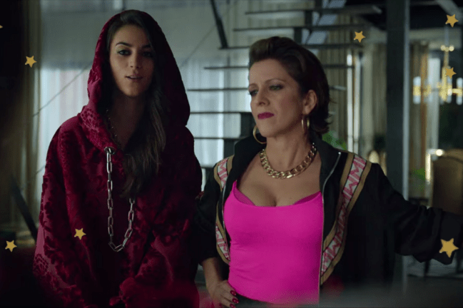 Looks quarta temporada Elite Foto com foco na Rebeca e sua mãe, do seriado Elite, Rebeca usa moletom vermelho e sua mãe blusa rosa pink. Elas estão em suas casas posando em frente a uma escada.
