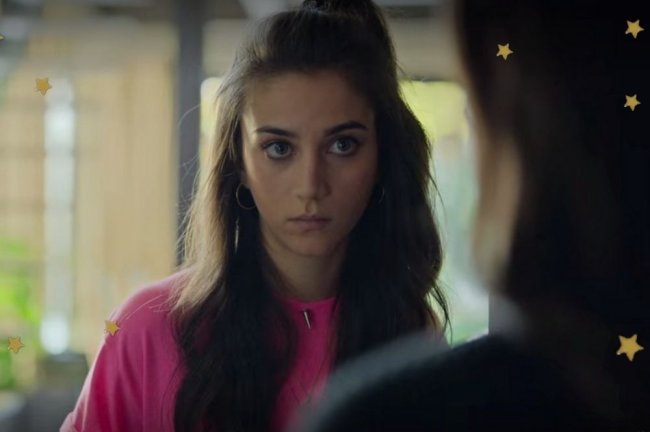 Foto com close no rosto da personagem Rebeca, usando blusa rosa, com penteado meio preso e expressão séria.