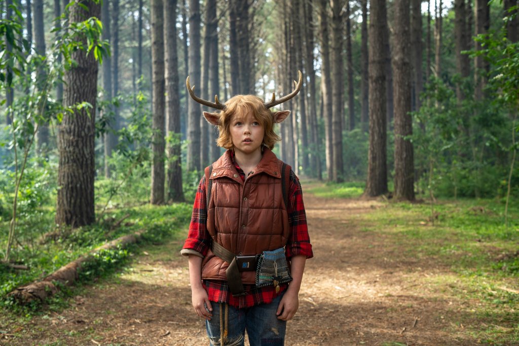 Cena do personagem Gus em Sweet Tooth; ele está no meio da floresta olhando assustado para frente e usa uma roupa vermelha desgastada com seus característicos chifres na cabeça