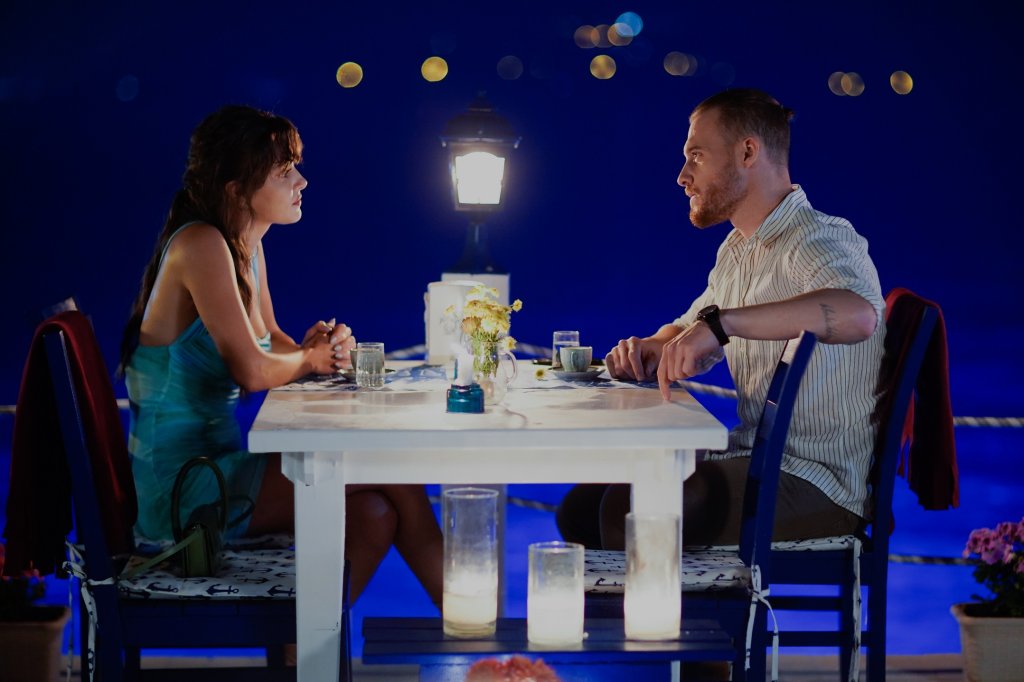 Eda e Serkan jantando; os dois estão sentados um de frente para o outro e se encaram; o céu azul da noite ilustra quase todo o fundo da imagem