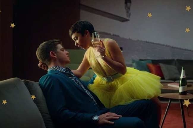Foto com foco nos personagens do seriado Elite, Guzman e Ari, sentados em sofá. Guzman usa terno e gravata e Ari vestido amarelo. Os dois se olham.
