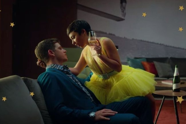 Vestido amarelo Ari Foto com foco nos personagens do seriado Elite, Guzman e Ari, sentados em sofá. Guzman usa terno e gravata e Ari vestido amarelo. Os dois se olham.