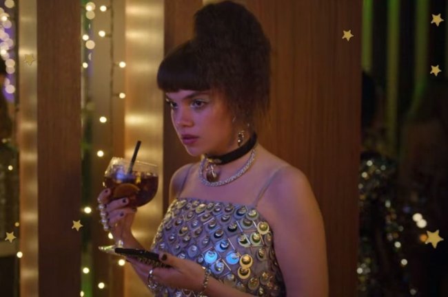 Foto com foco na personagem Mencía do seriado Elite, ela está segurando um copo com bebida, e um celular, ela usa chocker e vestido prata.