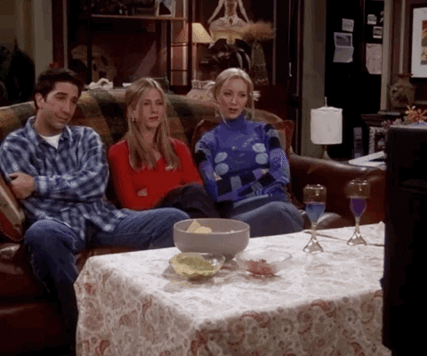 Ross, Rachel e Phoebe de Friends sentados no sofá assistindo TV e rindo