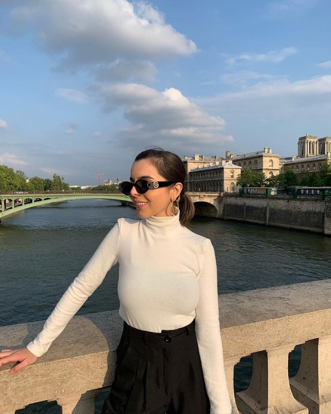 Foto de uma mulher em frente a um rio em Paris. Ela usa uma calça preta de alfaiataria, blusa branca de gola alta e manga longa. Olha para o lado e sorri.