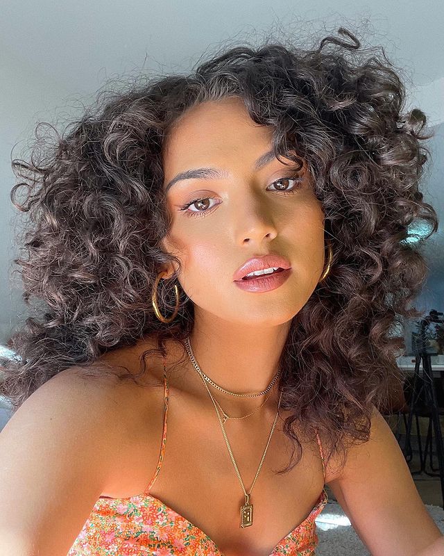 brinco de argola acessório clássico que combina com looks mais despojados Selfie de uma mulher. Ela usa um top de flores laranja, cabelo solto, colares pratas e brinco de argola dourado. Ela olha para a câmera e não sorri.
