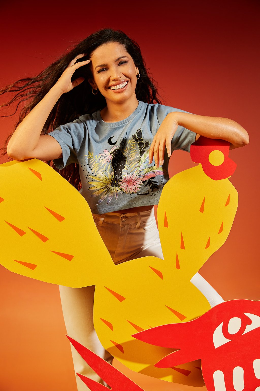 Juliette Freire usando uma camiseta azul cropped estampada com calça branca. Ela está sorrindo, com uma das mãos no cabelo e a outra apoiada em uma estrutura em formato de cacto amarelo e laranja.