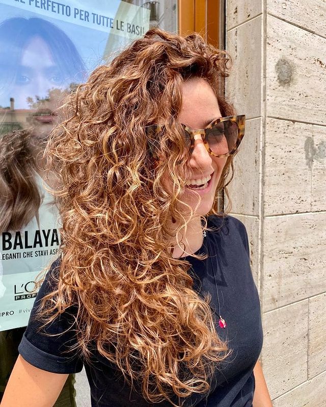 cabelo cacheado com smoky hair Jovem sorridente usando óculos escuros, com cabelos cacheados balançando com o vento e usando camiseta preta em frente a vidraça de um salão de cabeleireiro.