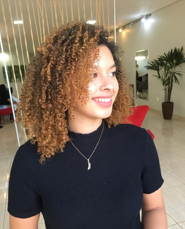 cabelo cacheado smoky Jovem sorridente olhando para o lado, posando em salão de cabeleireiro usando camiseta preta de gola alta com colar. Ainda na imagem conseguimos ver alguns detalhes do salão.