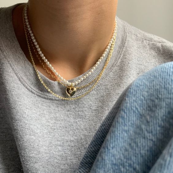 colar de pérolas é o acessório que deixa o look básico mais sofisticado Foto do pescoço de uma pessoa. Ela usa um colar de pérolas com pingente de coração, uma corrente dourada e blusa cinza.