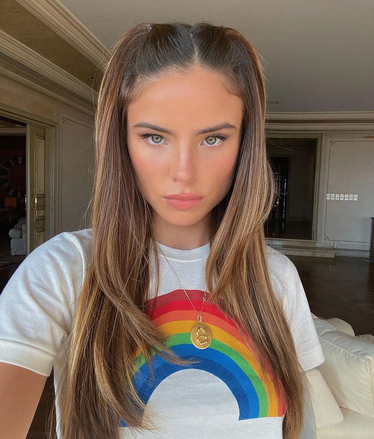 Selfie de Giulia Be; ela está olhando para câmera com uma blusa branca com desenho de arco-íris e expressão séria