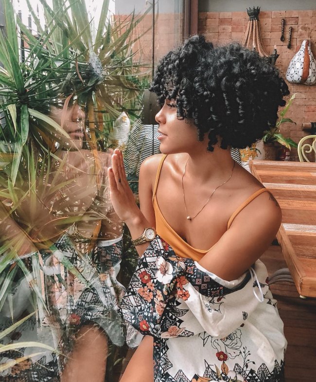 long-bob-cacheado-87 Jovem com expressão levemente sorridente, exibindo seu corte bob, usando regata laranja e olhando pelo vidro com uma das mãos apoiadas nele.