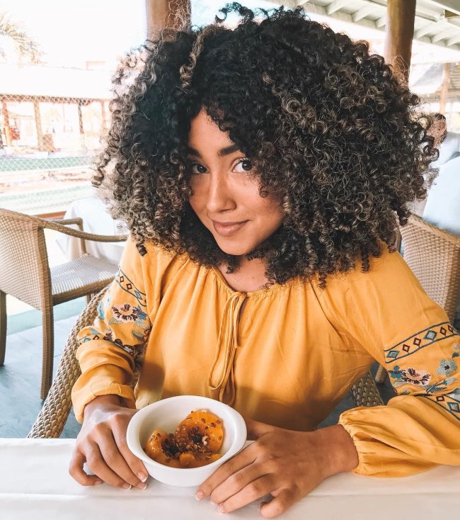 long-bob-cacheado-volumoso Jovem com expressão levemente sorridente, exibindo seu corte bob, usando blusa amarela e com potinho de frutas.