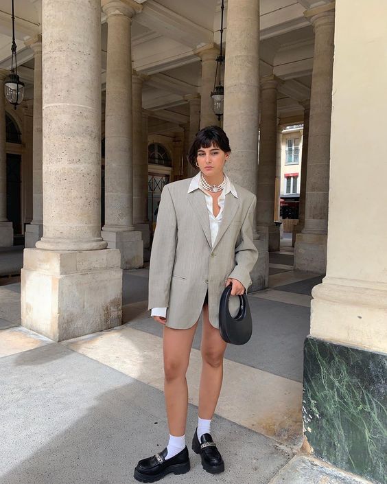 look com mocassim Jovem com expressão séria, posando em frente a colunas, usando blazer alongado cinza, camisa branca, e mocassim preto.