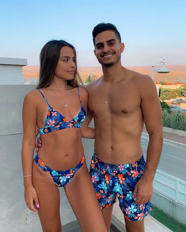 Casal abraçado com trajes de banho, a menina usa biquini azul com detalhes coloridos, e o menino usa bermuda com a mesma estampa do biquini dela. Ela aparece com o rosto virado para o lado direito e ele olha sorridente para frente.