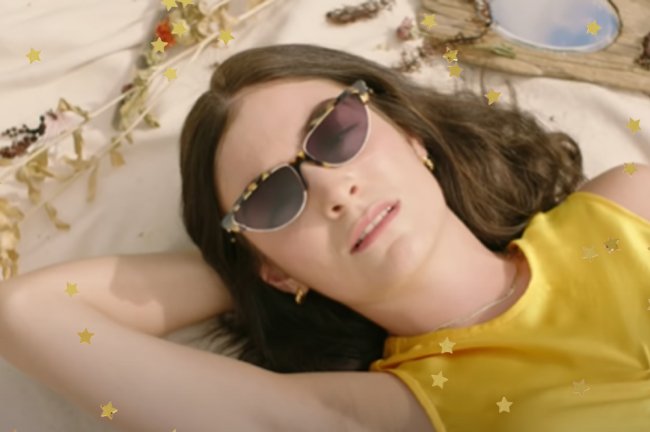 Frame do clipe de Solar Power de Lorde; a cantora está usando um cropped amarelo e usa um óculos de sol; ela está deitada com uma das mãos apoiadas atrás da cabeça e sorri levemente; estrelas amarelas decoram a imagem