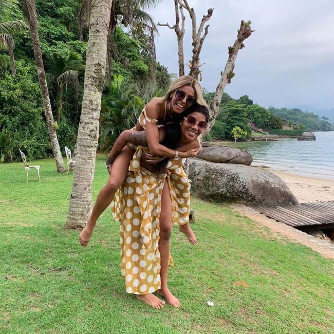 Ludmilla e Bruna posando em um local com grama e arvores. Bruna está montada nas costas de Ludmilla e as duas aparecem sorridentes.