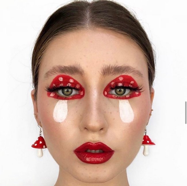 Foto do rosto de uma mulher em um fundo branco. Ela está com uma maquiagem em formato de cogumelo nos olhos, batom vermelho, cabelo preso em um coque baixo e brincos de cogumelo. Ela olha para a câmera e não sorri.