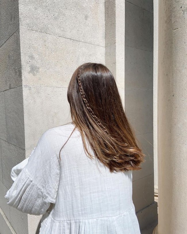 Foto de uma mulher de costas com uma mini trança no cabelo. Ela usa uma blusa branca e está com o cabelo liso solto.