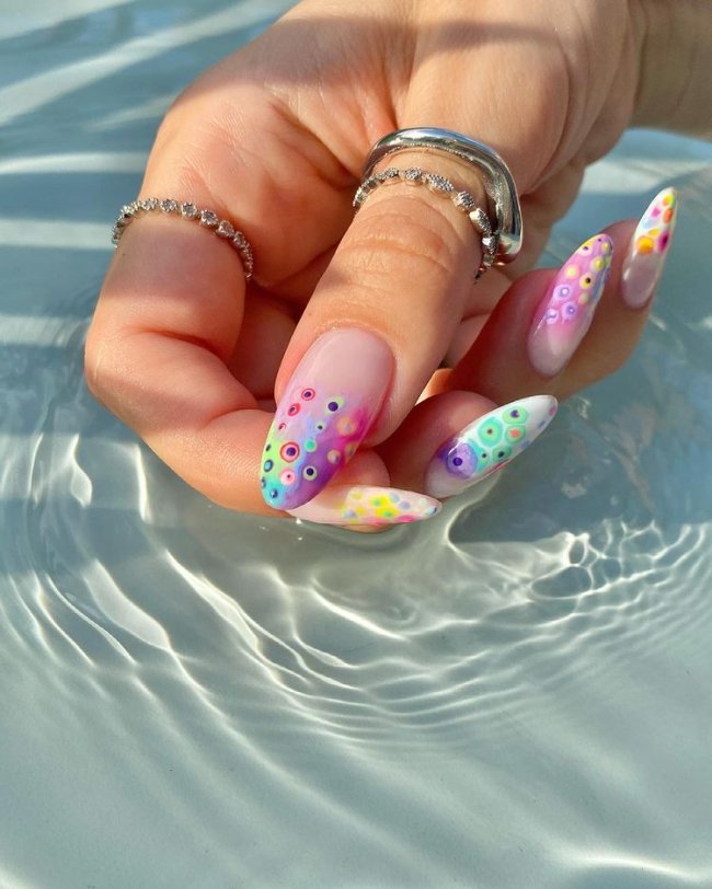 Foto com destaque nas unhas com nail art de bolinha, dessa vez com fundo transparente e várias bolinhas coloridas e irregulares na ponta da unha.