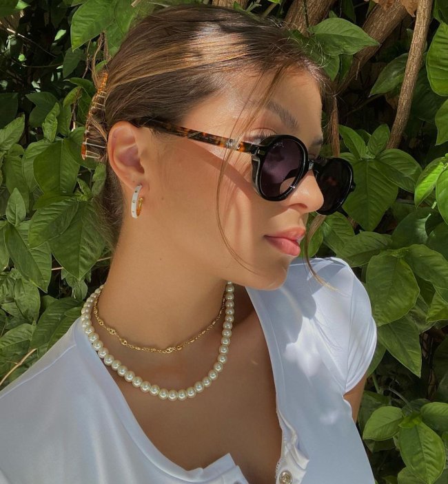 óculos de sol é o tipo de acessório que nunca sai de moda Selfie de uma garota com óculos de sol marrom de perfil. Ela usa uma blusa branca, colares, brinco de argola e cabelo preso em um coque.