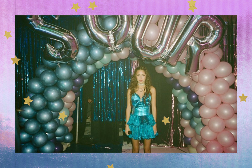 Olivia Rodrigo segurando um microfone na mão. Ao fundo balões azuis, rosa e lilás e com as letras que formam a palavra SOUR