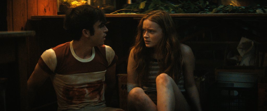 Ted Sutherland e Sadie Sink em cena de Rua do Medo parte 2: 1978; os dois estão sentados no chão; Sadie olha assustada para Ted que parece tentar observar algo atrás dela