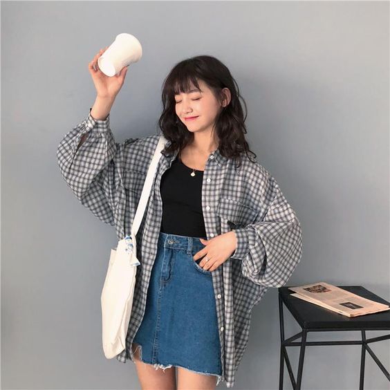 saia com camisa xadrez Foto de uma mulher. Ela usa saia jeans, blusa preta, camisa xadrez oversized e ecobag. Ela está segurando um copo de café na mão direita e sorri para a foto.