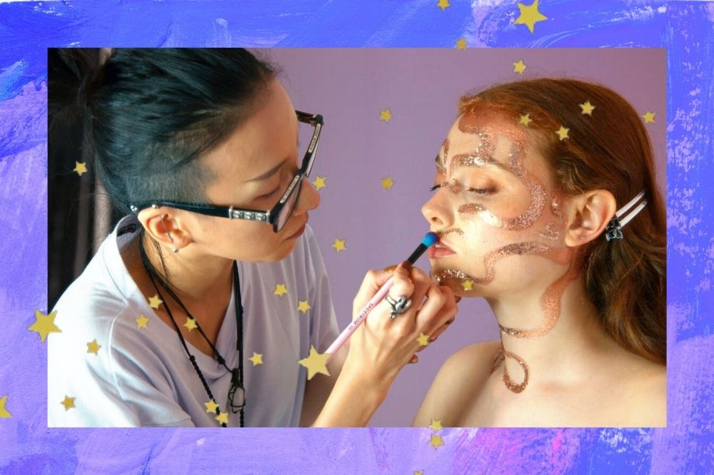 Montagem com fundo azul e estrelas douradas com a foto de uma cena do reality show Glow Up, da Netflix. Na foto, uma mulher com camiseta branca, óculos e cabelo preso faz a maquiagem de uma modelo com cabelo ruivo solto.