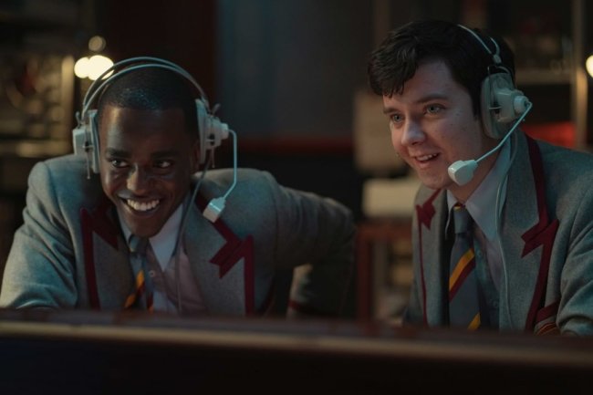 Otis e Eric usando o uniforme da escola e olhando para uma tela de computador enquanto usam fones de ouvido com microfone; os dois estão sorrindo e parecem animados com o que estão assistindo