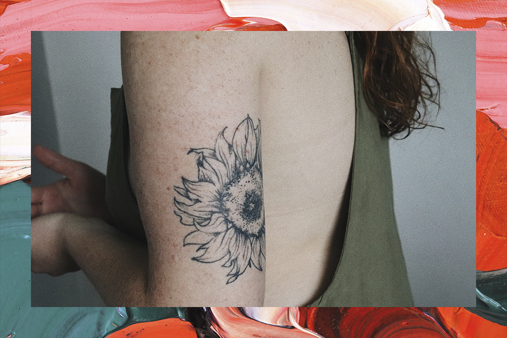 Na imagem, aparece o tronco de uma mulher que veste uma camiseta verde sem mangas.. Em seu braço está tatuado uma flor.