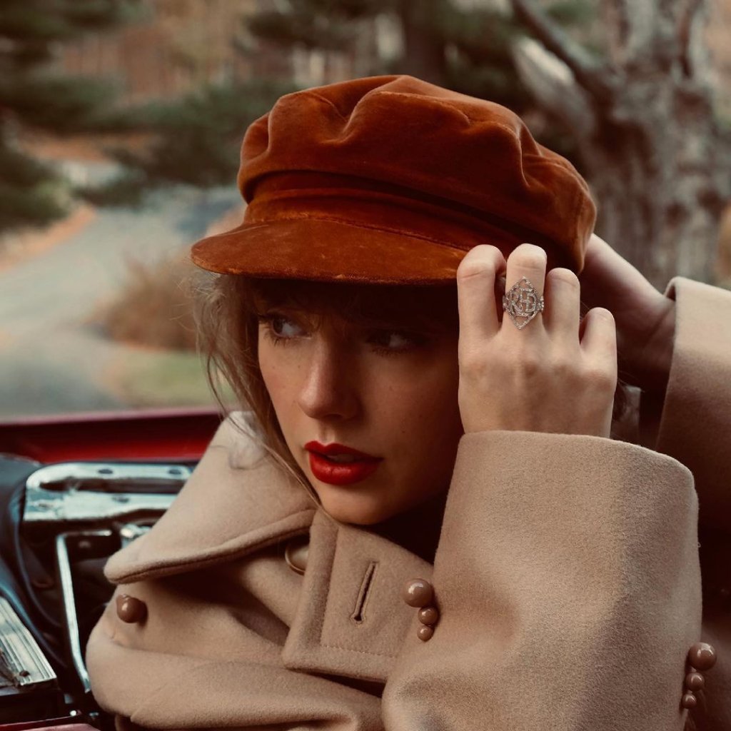 Taylor Swift usando uma boina e olhando para o lado; ela está de batom vermelho e com um casaco bege