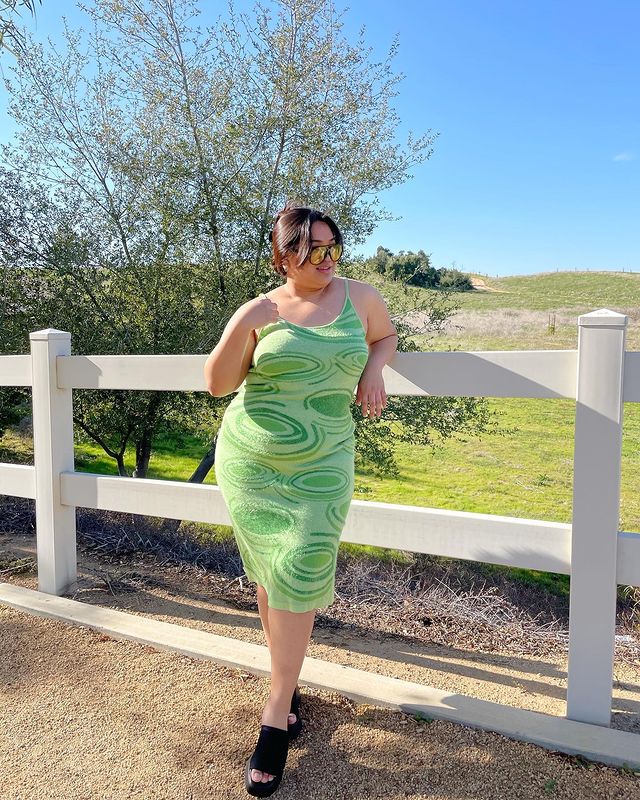 Foto de uma mulher em frente a uma cerca. Ela usa um vestido verde menta com estampa oval em tons de um verde mais escuro, óculos de sol com lente amarela, tamanco preto e cabelo preso em um coque. Ela olha para o lado e sorri levemente.