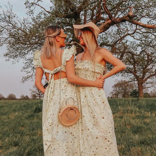 Casal posa em frente a arvores em chão com gramado, as duas estão se olhando e o cabelo cobre suas expressões faciais. Uma está de lado para a câmera e a outra de costas. As duas usam vestido com estampa floral igual.