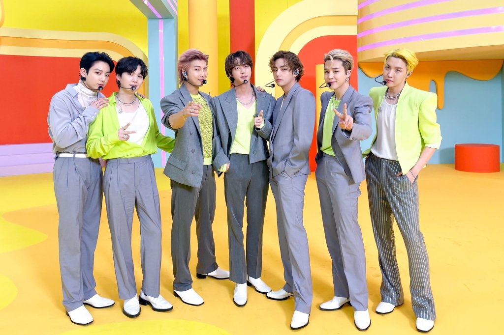Integrantes do BTS em imagem de divulgação em um fundo amarelo com detalhes coloridos; eles estão alinhados usando roupas em tons de azul claro, verde, branco e cinza escuro