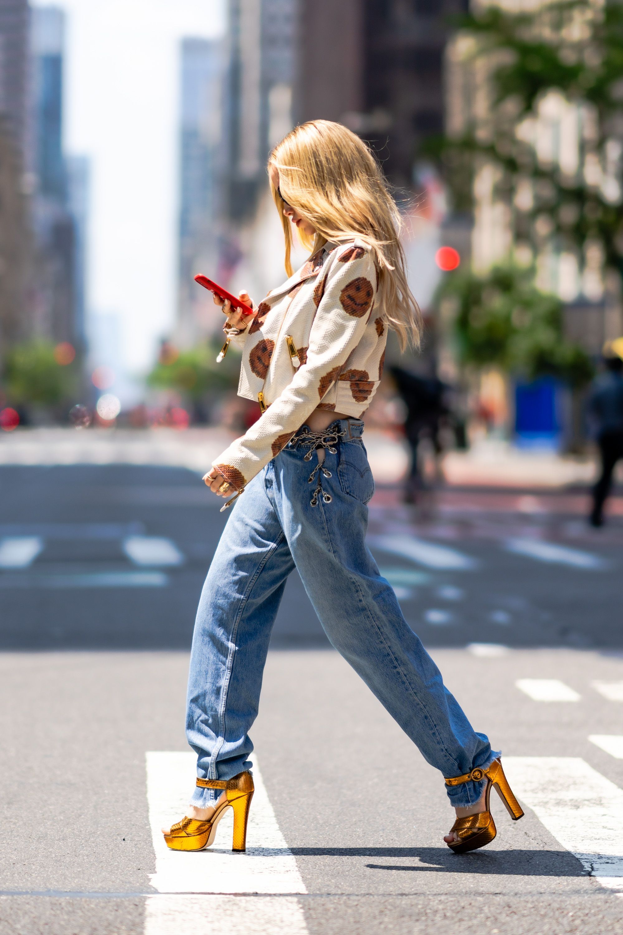 Foto da atriz e cantora Dove Cameron de lado, caminhando em uma rua. Ela está olhando para seu celular, usando jaqueta e calça jeans.