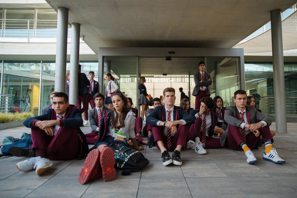Omar, Rebeka, Guzmán, Samuel e Ander em cena de Elite; eles estão sentados no chão na porta da escola junto com outros alunos, todos uniformizados, em uma espécie de protesto