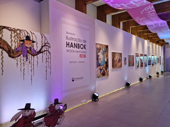 Exposição-Woo NaYoung Imagem da Exposição Woo NaYoung; uma parede branca com luzes em tons de rosa/roxo iluminam as artes expostas