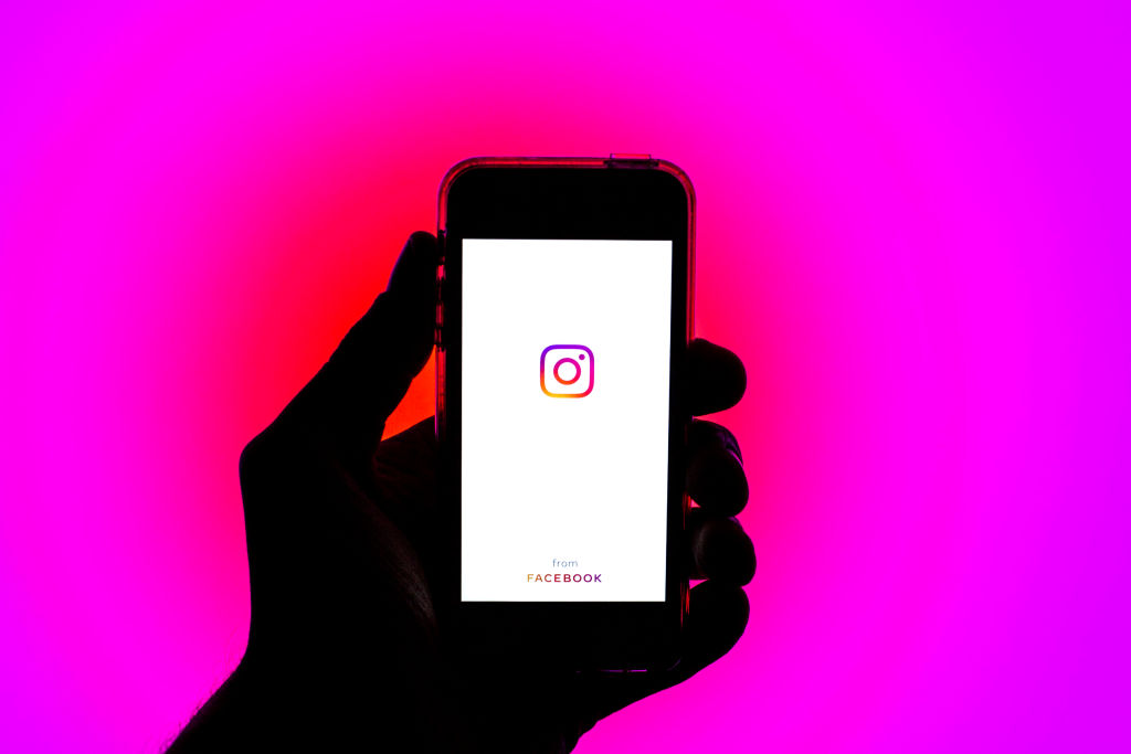 Foto de uma mão segurando um celular apresentando o logo do Instagram. Fundo é pink.