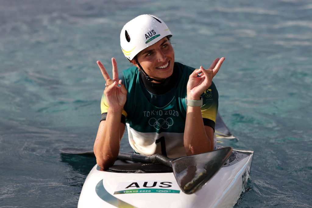 A australiana Jessica Fox comemorando seu feito em Tóquio. Ela está de capacete branco, sentada em uma caiaque, no mar, fazendo sinal de paz e amor com as mãos