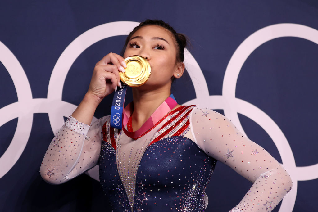 A hmong Sunisa Lee, dos EUA, beijando sua medalha de ouro após vencer os individuais gerais da ginástica; ela está com uma das mãos na cintura e veste um collant azul, branco e vermelho bastante brilhante