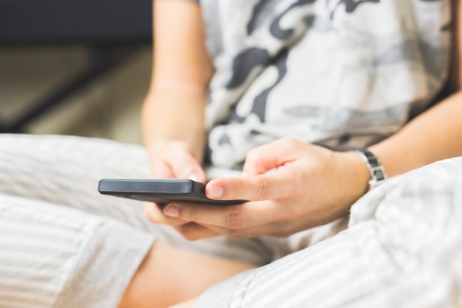 Uma adolescentes sentada na cama de pernas cruzadas mexendo no celular. O rosto da jovem não aparece.