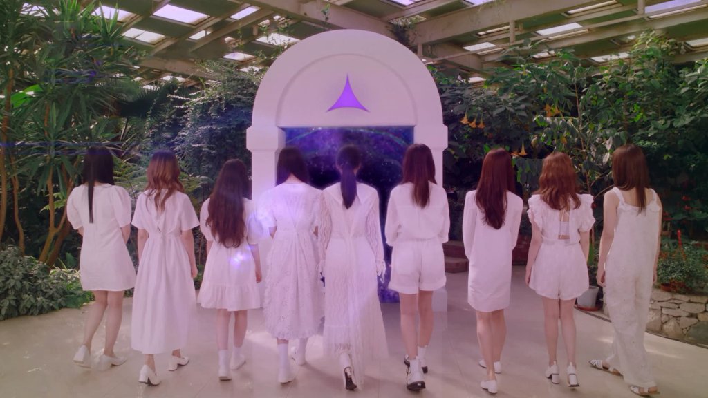 Girls Planet 999 com diversas integrantes de costas para a câmera usando roupas brancas na frente de uma espécie de portal branco com detalhe lilás; a iluminação também está em tons de roxo